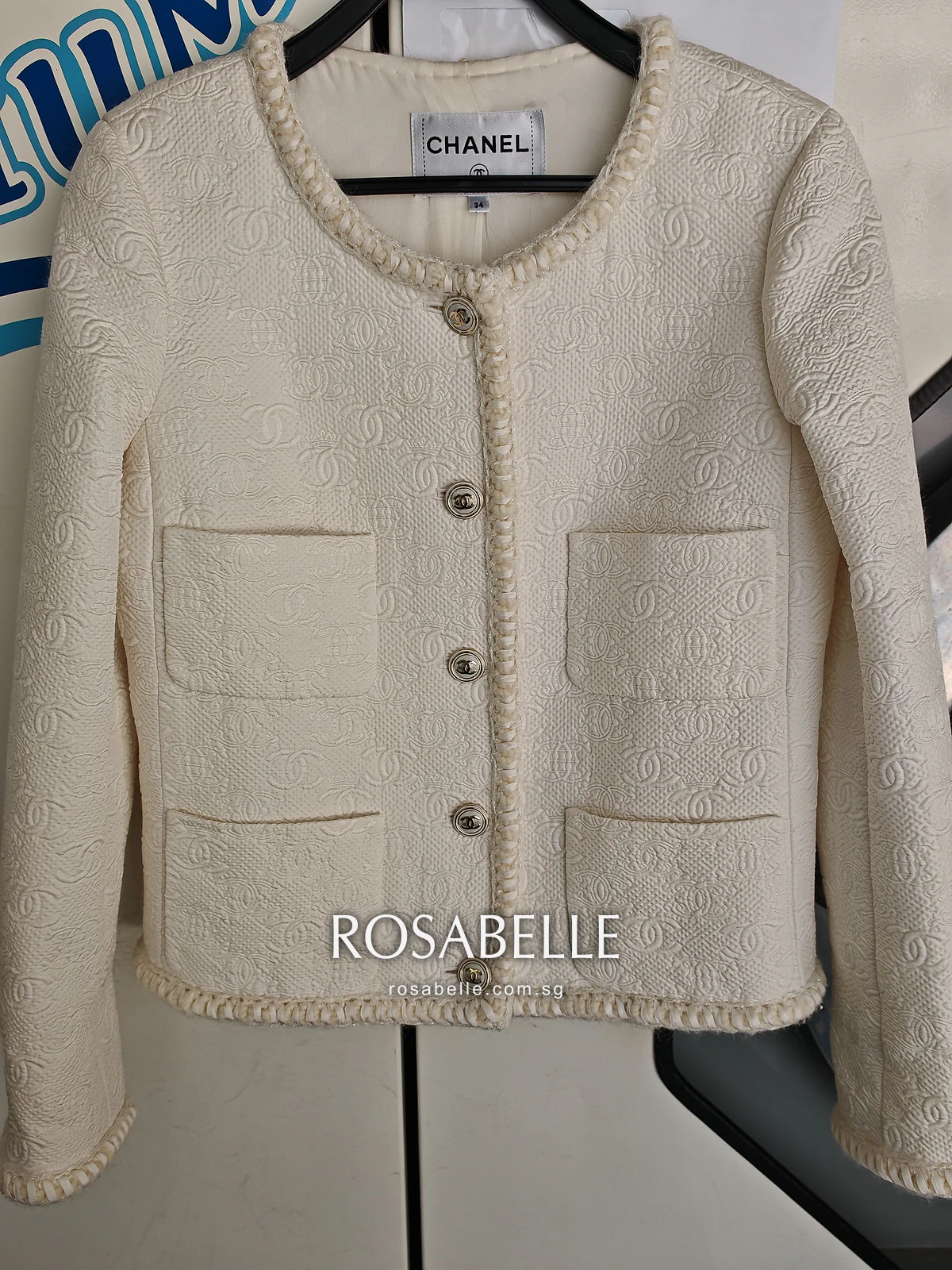 The cleaned cream Chanel jacket on a hanger against a pale wall, four patch pockets and braided edges in even light