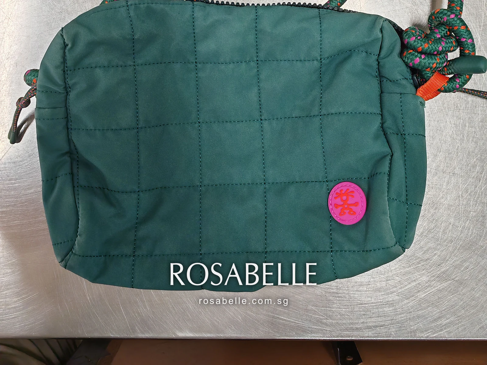 Teal Crumpler bag with grid quilting and a round pink badge, lying on a steel bench before cleaning