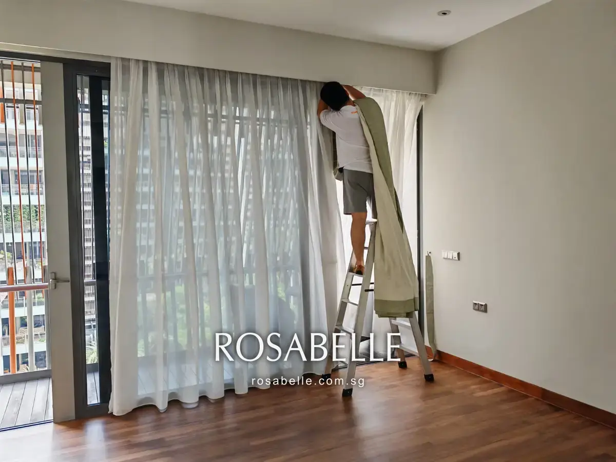 The green curtain panel folded over the shoulder of a Rosabelle Launderette staff member on a stepladder, with the white sheer left drawn across the window