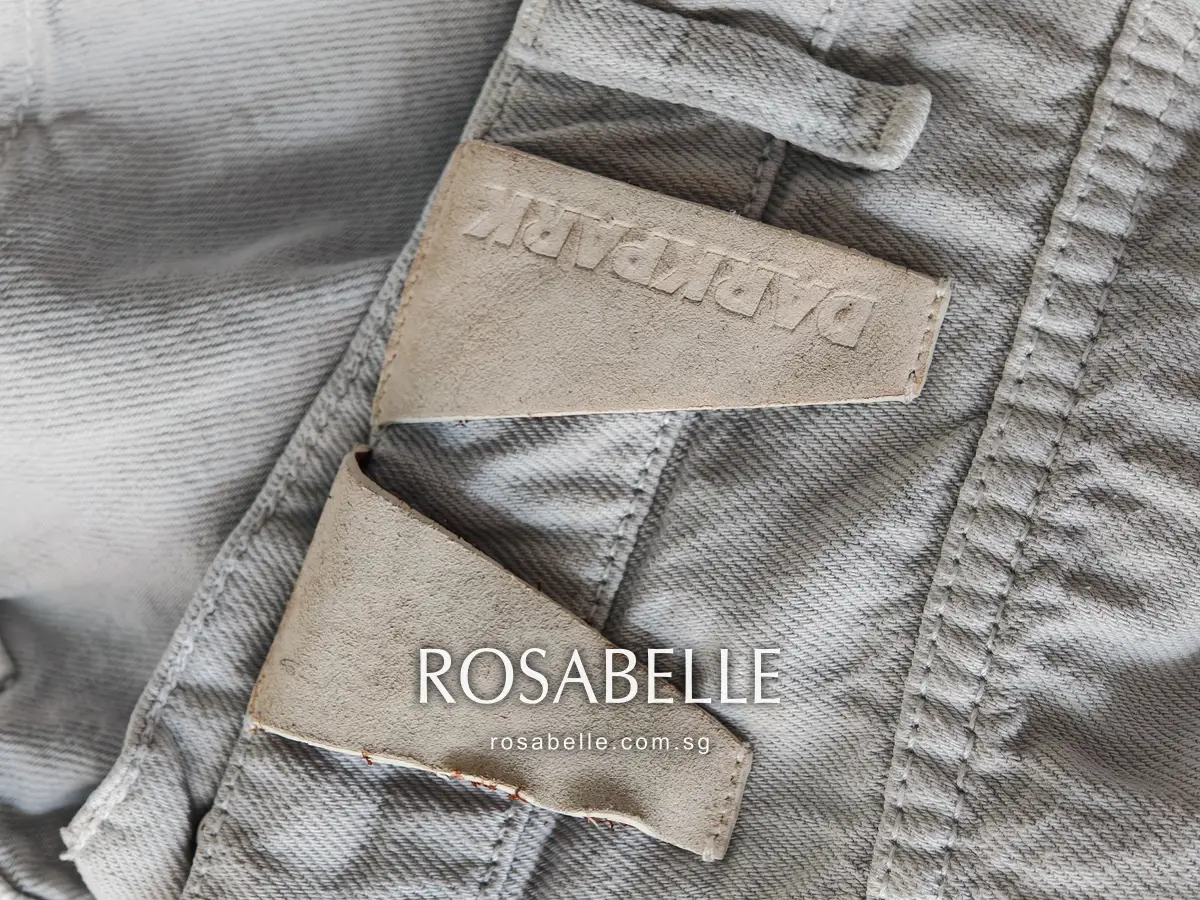 Debossed Darkpark leather back patch in pale beige cut in two pieces, sitting on grey denim beside a belt loop