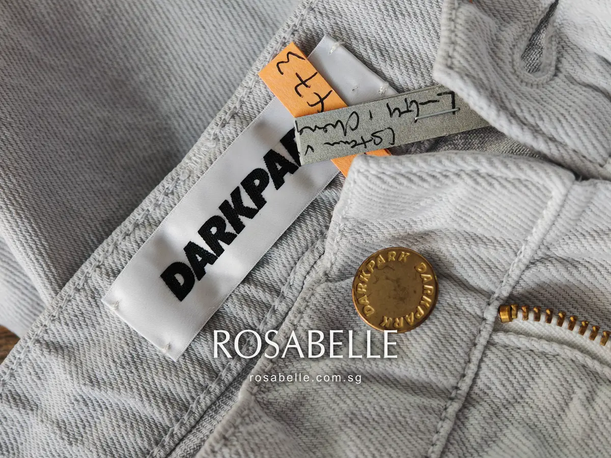 White woven Darkpark label and gold shank button on grey-tinted denim twill, a shop ticket pinned at the waistband