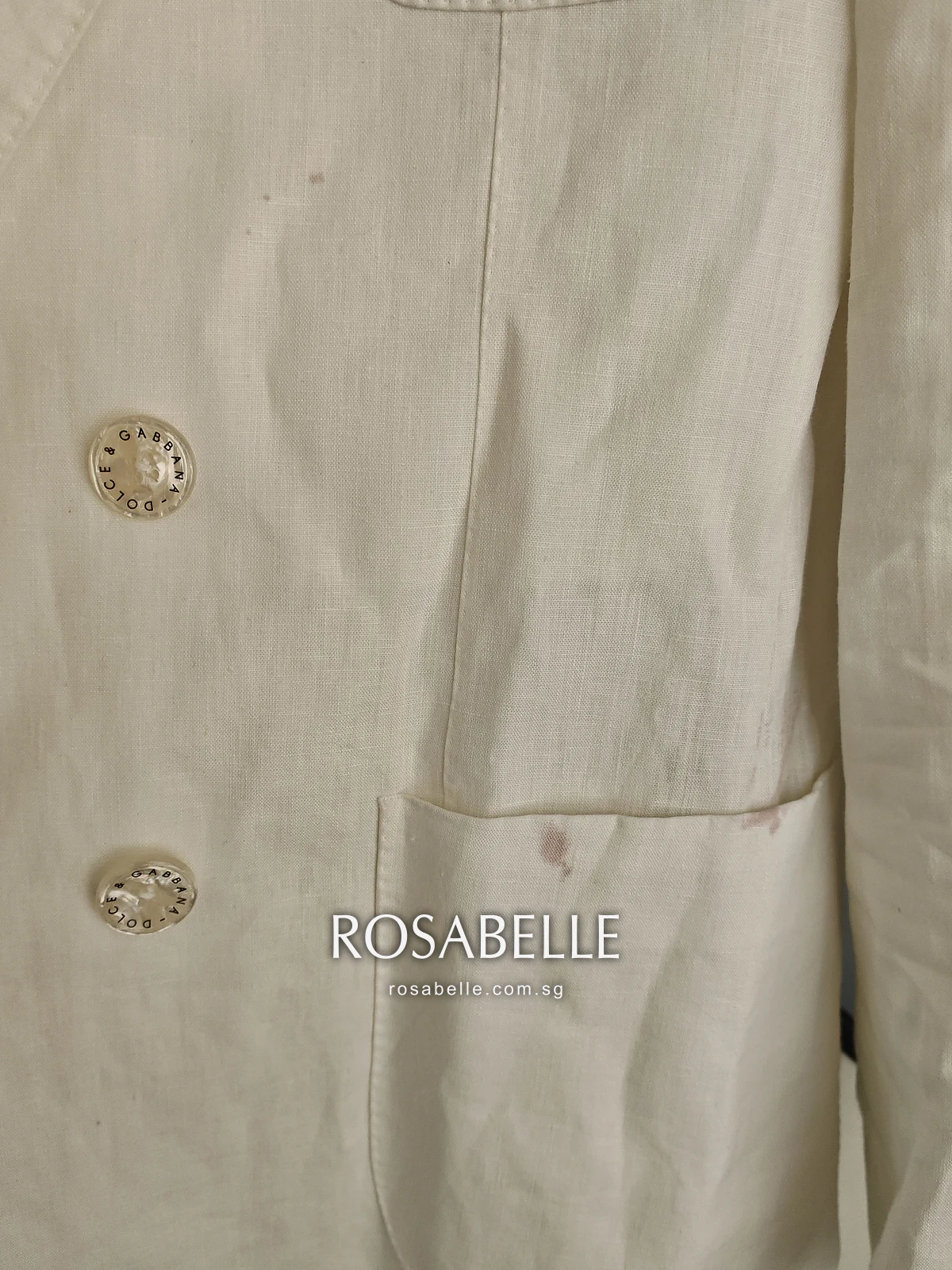 Close view of the linen coat front, small reddish wine spots above the patch pocket beside two logo buttons