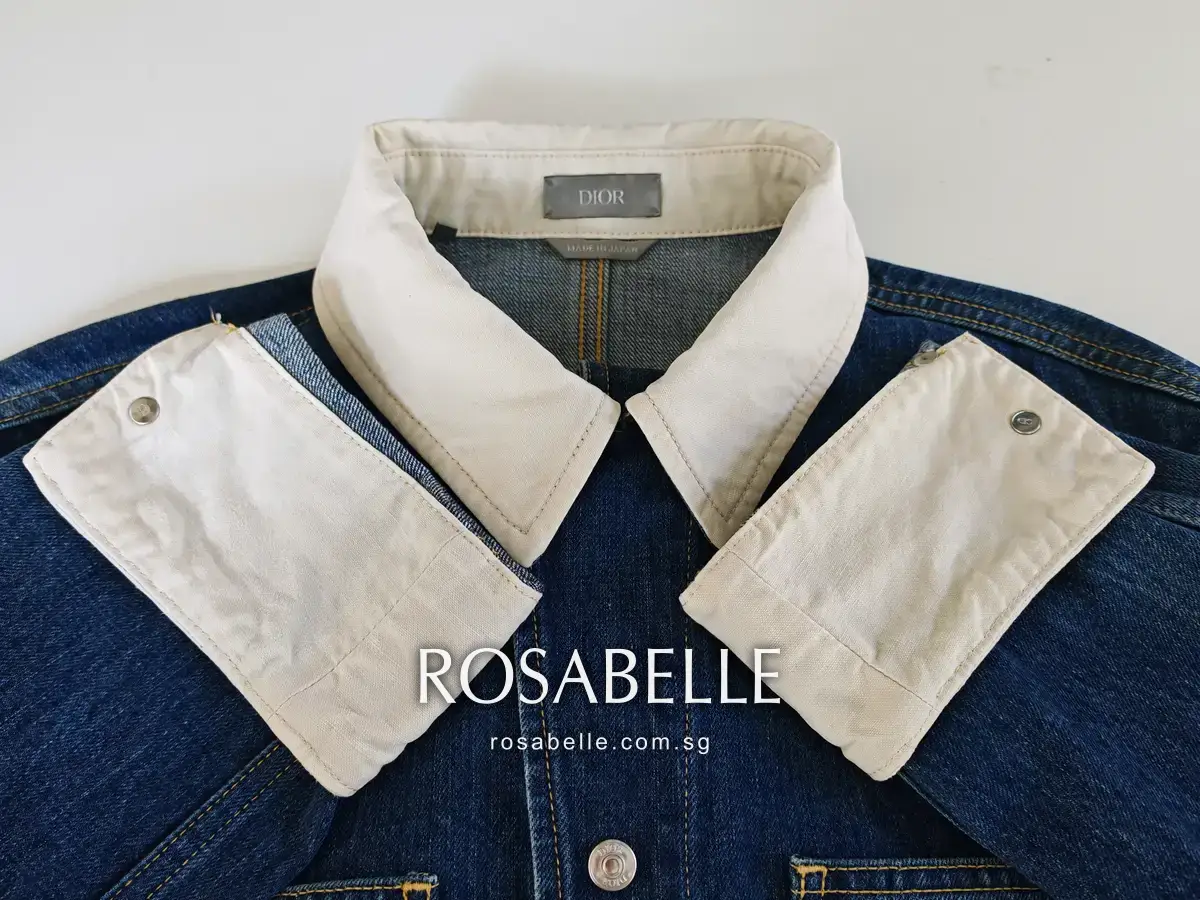 The cleaned Dior denim jacket collar standing with both cuff facings folded out, the cream cotton an even colour with no speckling, photographed against the same white wall as the before frame