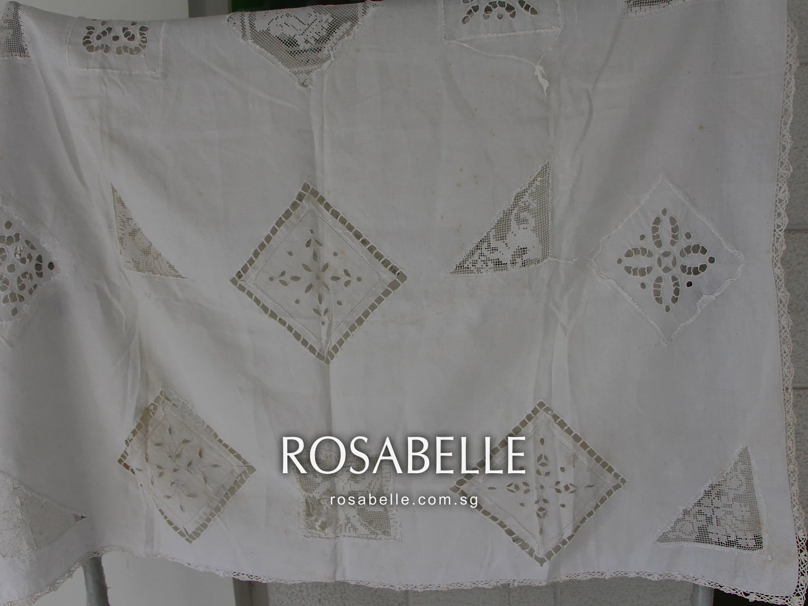 Linen tablecloth with cutwork diamonds and lace insets hung on a rack, brown age staining across its lower half