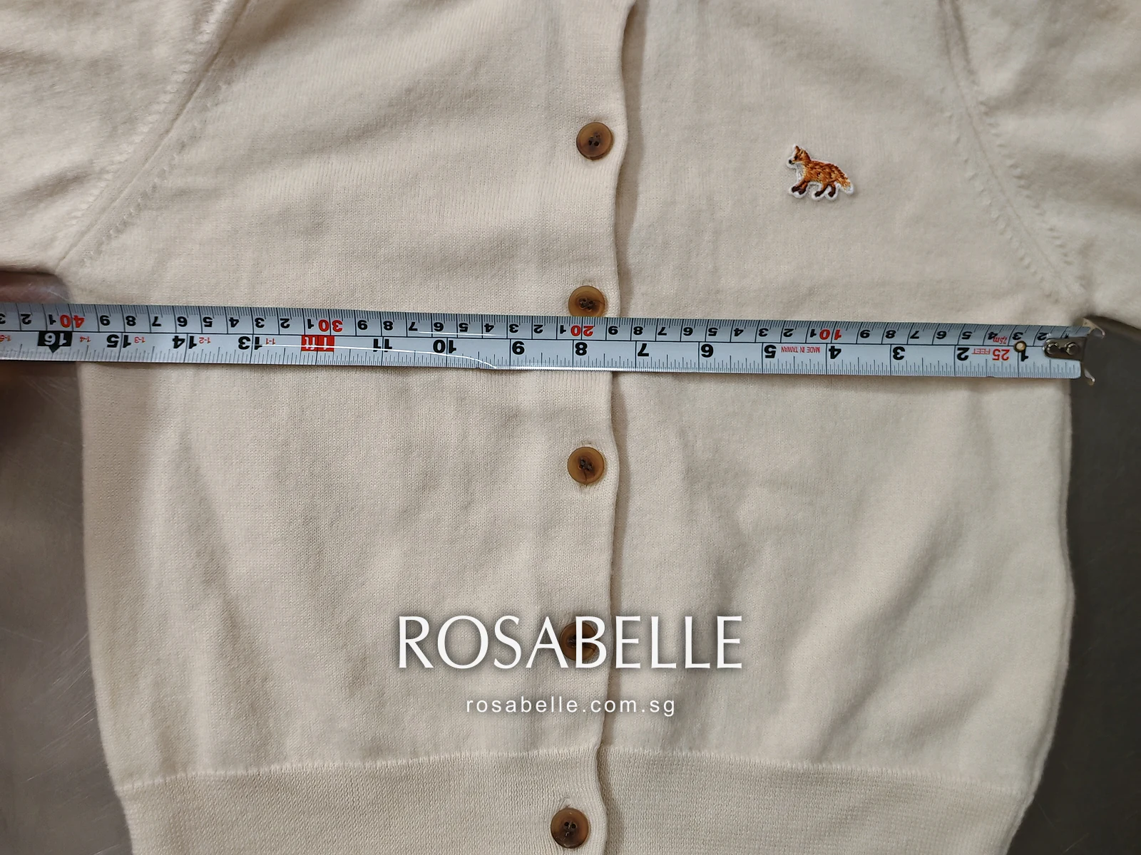 Tape measure laid across the chest of the shrunken wool cardigan just below the armholes, fox patch above it