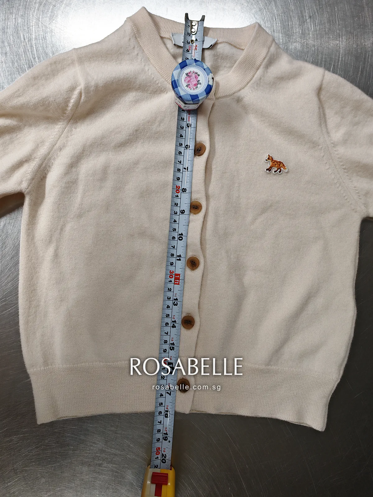 Beige Maison Kitsuné cardigan laid flat on a steel table, a tape measure running from collar to hem along the buttons