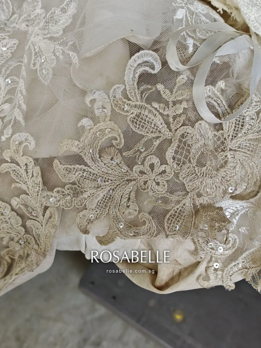 Beige-tinged lace appliqués at the gown hem with grubby satin folded behind, soil caught in the corded outlines