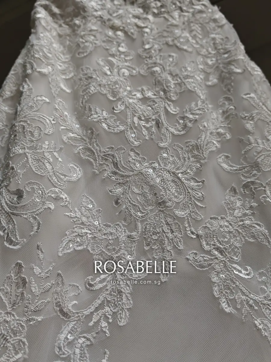 Dense white floral lace across the mermaid skirt, small sequins catching the light among the leaves
