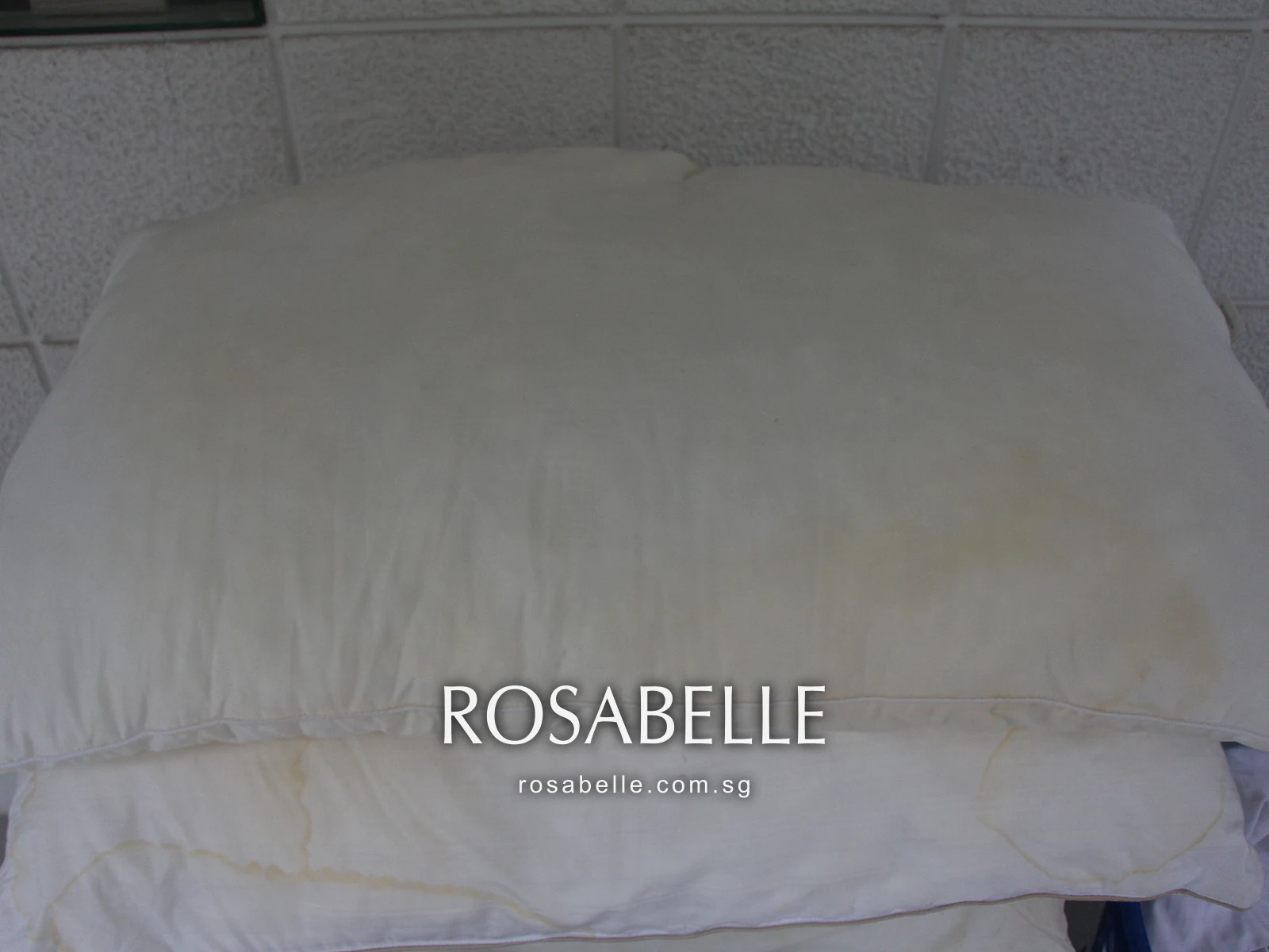 Plain long white pillow with faint yellow tide marks across its face, stacked on a second marked pillow